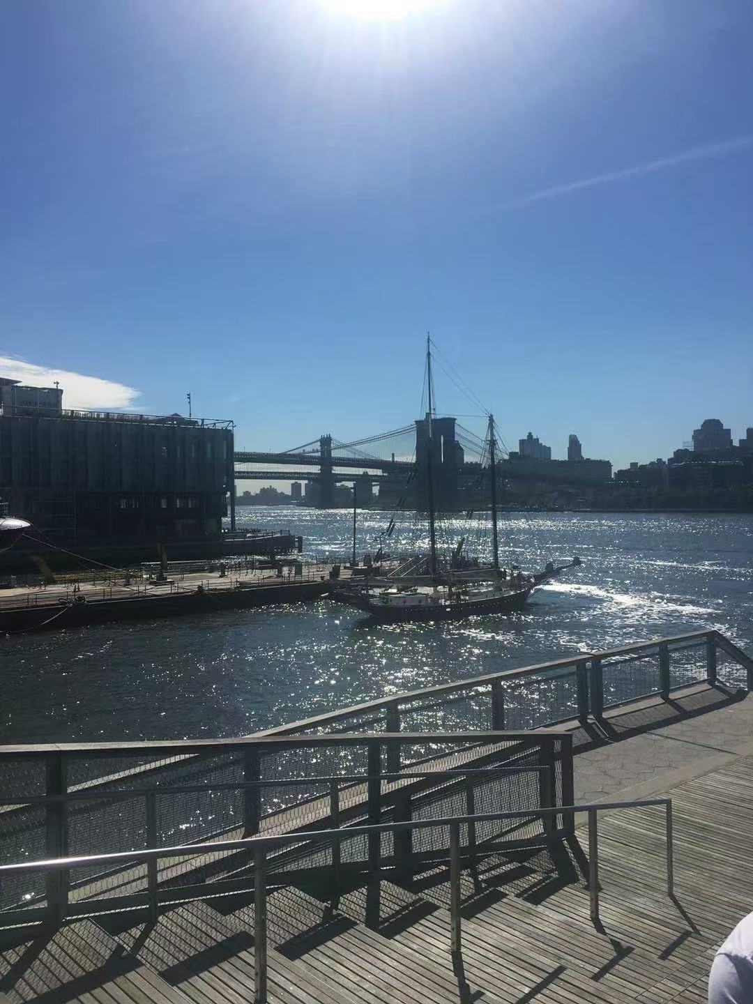 A sailboat on a sunlit waterfront with a suspension bridge in the background.