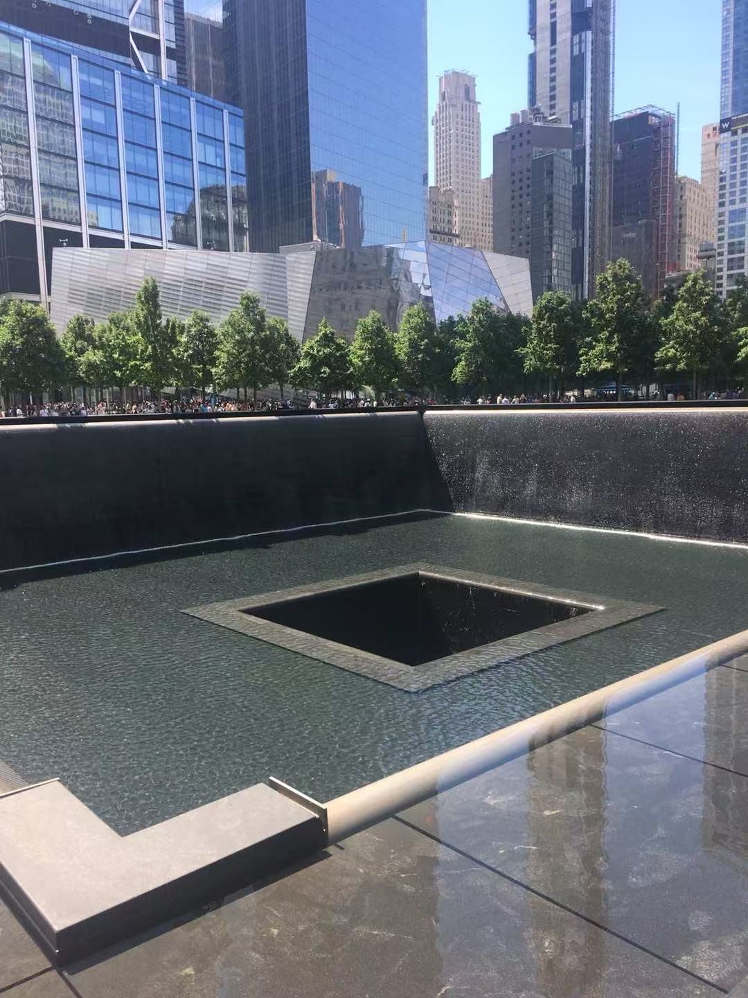A square memorial reflecting pool surrounded by trees and city buildings.